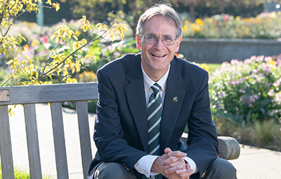 Dean Eric L. Hegg sitting at a bench with his right elbow on the back while he is turned facing the camera and smiling.