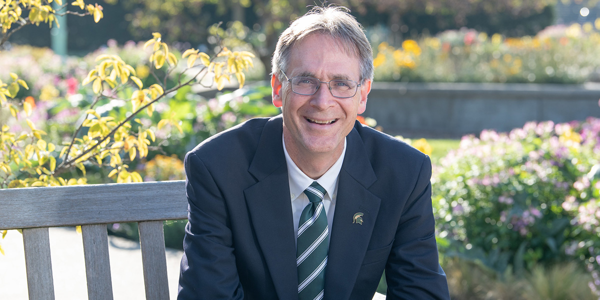 Dean Eric L. Hegg sitting at a bench with his right elbow on the back while he is turned facing the camera and smiling.