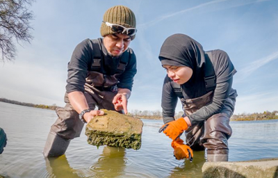 Two students, one male the other female, working in the water. The male student is holding a rock.